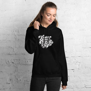 AG - Woman wearing a black hoodie with, know your worth, text against a white brick wall.