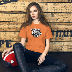 AG - Woman wearing a burnt orange t-shirt, with know your worth text, sitting against a wooden wall.