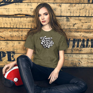 AG - Woman wearing a heather olive t-shirt, with know your worth text, sitting against a wooden wall.
