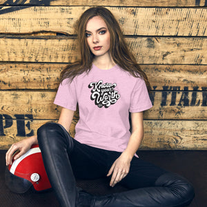 AG - Woman wearing a heather prism t-shirt, with know your worth text, sitting against a wooden wall.