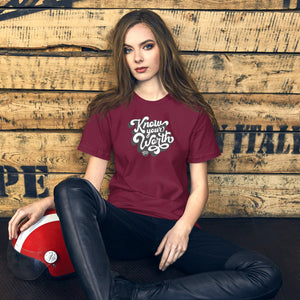 AG - Woman wearing a maroon t-shirt, with know your worth text, sitting against a wooden wall.