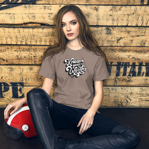 AG - Woman wearing a pebble t-shirt, with know your worth text, sitting against a wooden wall.