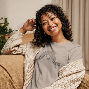 AG - Woman wearing a gray t-shirt with a musical graphic design, sitting on a couch in a cozy indoor setting.